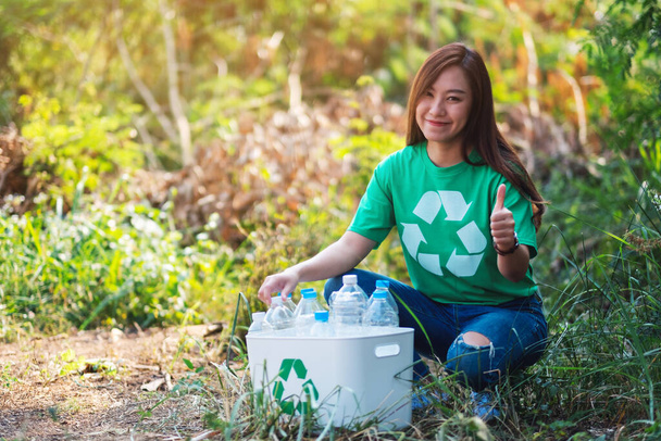 Eine schöne asiatische Frau macht Daumen nach oben Handzeichen beim Sammeln von Müll Plastikflaschen in einen Papierkorb in der Natur - Foto, Bild