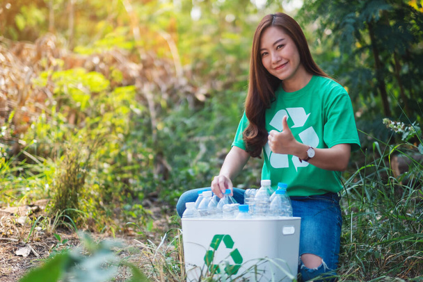 屋外のごみ箱にゴミプラスチックボトルを収集しながら、親指を作る美しいアジアの女性 - 写真・画像