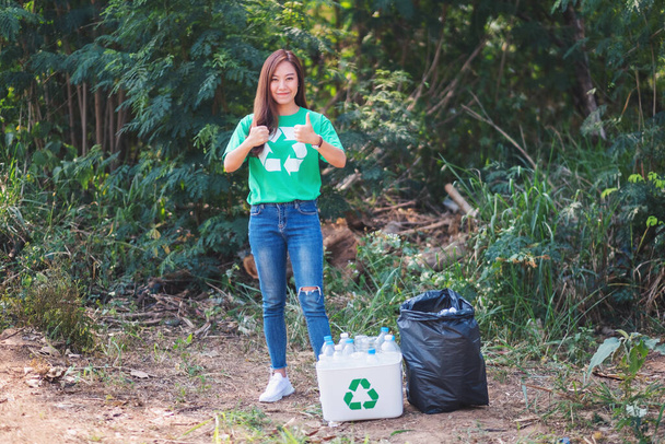 Una hermosa mujer asiática haciendo pulgares hacia arriba signo de mano mientras recoge botellas de plástico de basura en una papelera de reciclaje en el aire libre - Foto, imagen
