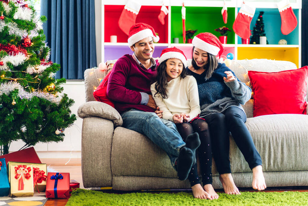 幸せなアジアの家族の父親と母親の肖像とサンタの帽子の娘は、カメラを見て、クリスマスの時間に家で一緒に過ごすことを楽しんでいます - 写真・画像