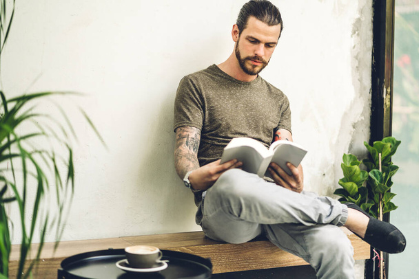 Όμορφος hipster άνθρωπος χαλαρώνοντας ποτό καφέ και να διαβάσετε το βιβλίο εργασίας μελέτη και κοιτάζοντας το περιοδικό σελίδα, ενώ κάθεται στην καρέκλα στο καφέ και το εστιατόριο - Φωτογραφία, εικόνα