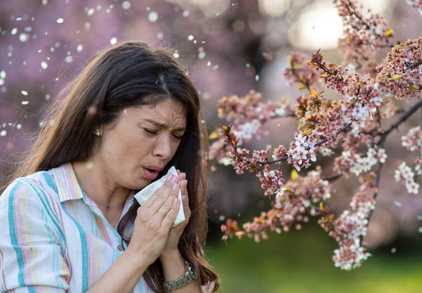 Afraid young woman sneezing in napkin in front of blooming tree. Spring allergy attack concept - Foto, Bild