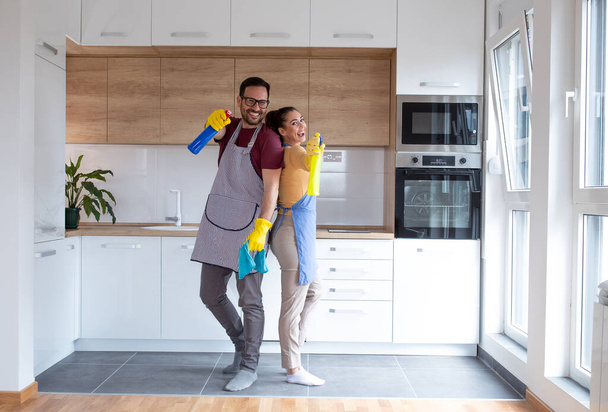 Νεαρό ζευγάρι στην κουζίνα με είδη καθαρισμού φορώντας γάντια και κάνοντας τις δουλειές τους μαζί. - Φωτογραφία, εικόνα