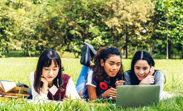 Gruppe von lächelnden internationalen Studenten oder Jugendlichen, die mit Laptop sitzen und lernen, während sie ihre Hausaufgaben machen und gemeinsam im Park der Universität Bücher lesen. - Foto, Bild