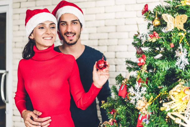 Romantic sweet couple in santa hats having fun decorating christmas tree and smiling while celebrating new year eve and enjoying spending time together in christmas time at home - Foto, Bild