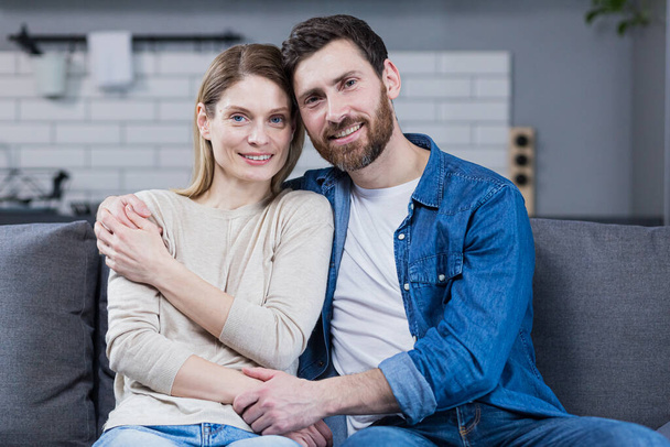 Young married couple man and woman sitting together on sofa and hugging, portrait of middle aged family looking at camera - Foto, Imagem
