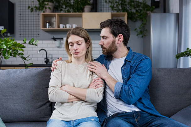 A man tries to support and calm a sad woman, a married couple sitting on a sofa at home - Fotoğraf, Görsel