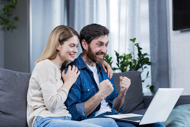 Happy couple man and woman looking at laptop screen, celebrating victory, holding hands up, gesture of victory and success - Fotoğraf, Görsel
