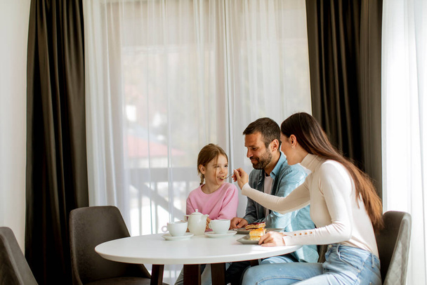 Ευτυχισμένη νεαρή οικογένεια τρώνε κέικ και πίνουν τσάι μαζί στο δωμάτιο - Φωτογραφία, εικόνα