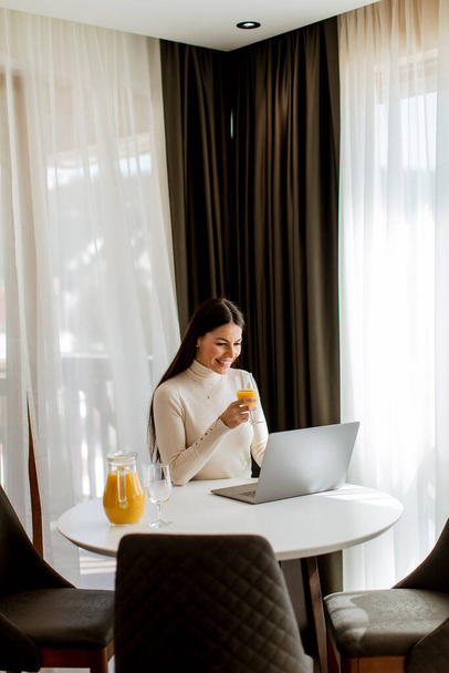 Молода жінка використовує ноутбук і п'є здоровий апельсиновий сік у кімнаті
 - Фото, зображення