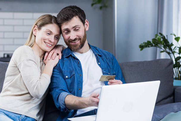Happy married couple man and woman sitting at home on the couch using laptop for online shopping, holding a credit bank card - Photo, image
