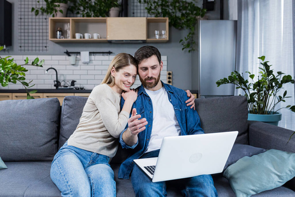 Married couple man and woman sitting together at home on the couch using laptop for video chat and chatting with friends - Foto, Imagen