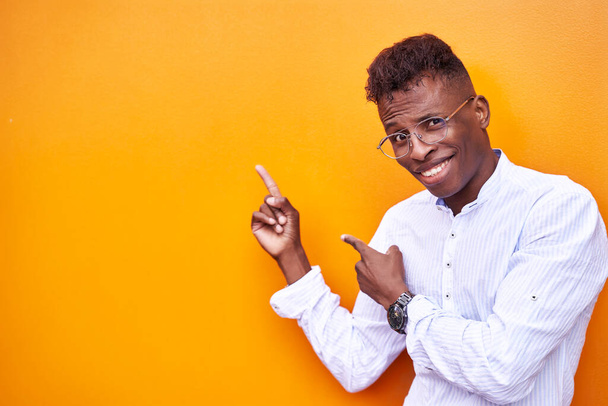 Hombre de negocios afroamericano con camisa inteligente y gafas sobre fondo naranja aislado sonriendo y mirando a la cámara apuntando con las manos y los dedos hacia un lado - Foto, Imagen