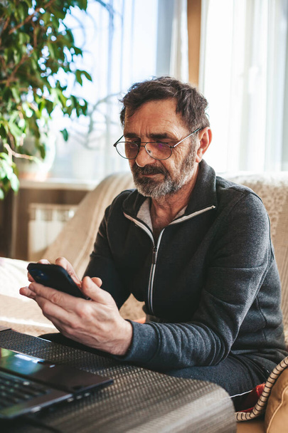 Serious mature man in glasses sitting on a couch texting or messaging or searching or reading on his smartphone. Seniors using technology concept. - Photo, image