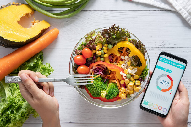 Calories counting , diet , food control and weight loss concept. Calorie counter application on smartphone screen at dining table with salad, fruit juice, bread and fresh vegetable. healthy eating - 写真・画像
