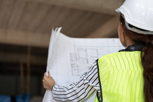 Asijský inženýr nebo mladá žena Architekt nasadit přilbu pro bezpečnost a podívat se na plán pro inspekci stavební továrny staveniště na staveništi. Inteligentní pracující žena koncept. - Fotografie, Obrázek