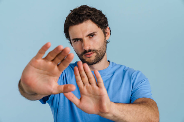 Young brunette man with bristle showing stop gesture at camera isolated over blue background - Foto, Bild