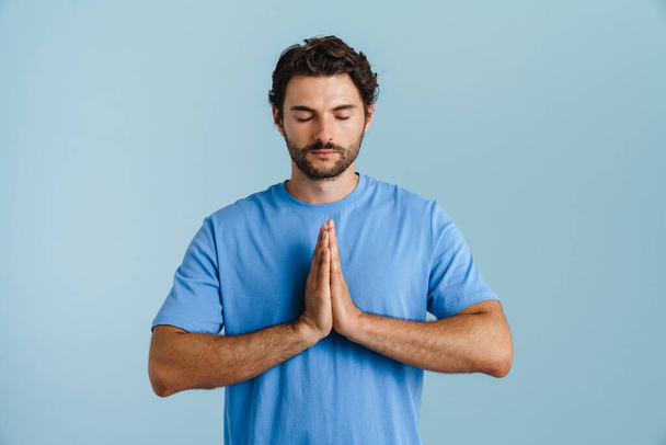 Young brunette man with bristle gesturing while meditating isolated over blue background - Foto, imagen