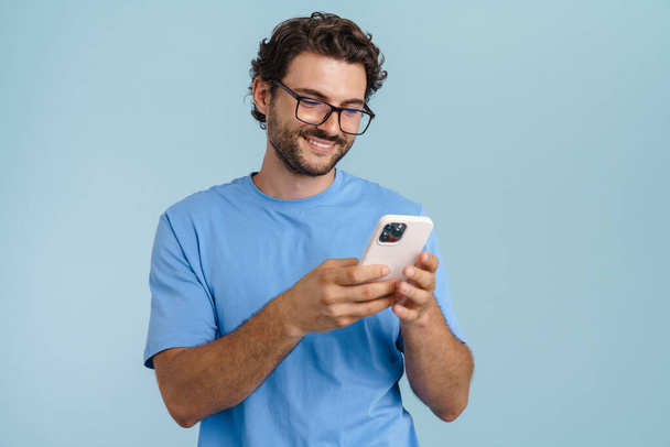 Young brunette man smiling while using mobile phone isolated over blue background - Zdjęcie, obraz