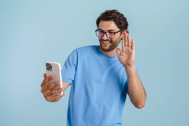 Young brunette man gesturing while using mobile phone isolated over blue background - Foto, Imagen
