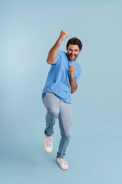 Young brunette man screaming while making winner gesture isolated over blue background - Fotoğraf, Görsel
