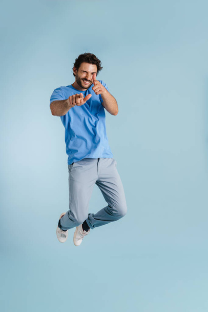 Young brunette man jumping while pointing fingers at camera isolated over blue background - Fotó, kép