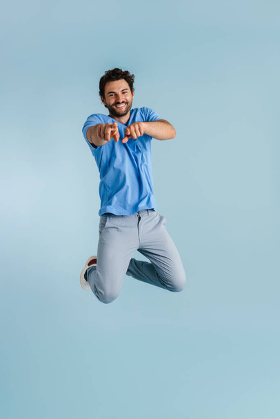 Young brunette man jumping while pointing fingers at camera isolated over blue background - Фото, изображение