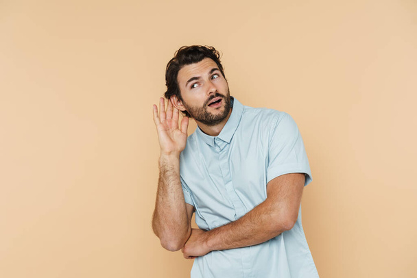 Young white man gesturing and listening while looking aside isolated over beige background - Foto, Imagem