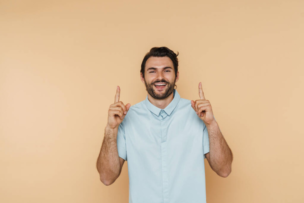 White bristle man wearing shirt smiling and pointing fingers upward isolated over beige background - Photo, Image
