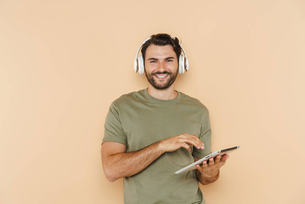 White bristle man using tablet computer and headphones isolated over beige background - 写真・画像