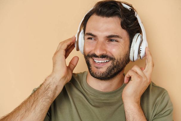 Young white man smiling while listening music with headphones isolated over beige background - Photo, image