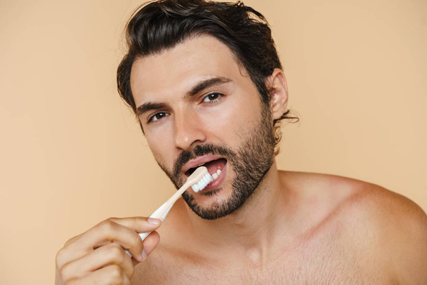 Young half-naked man looking at camera while brushing his teeth isolated over beige background - Φωτογραφία, εικόνα