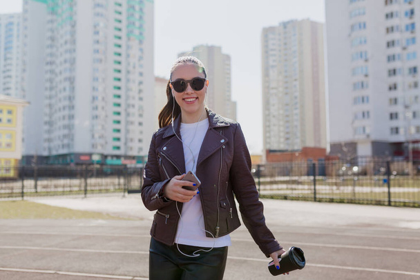 Positive stylish lady with collected dark hair in sunglasses and burgundy jacket smiling and listening music on city background - Фото, изображение