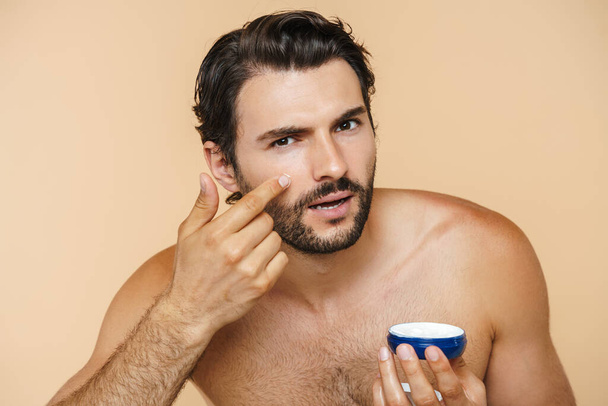 Young half-naked man looking at camera while applying face cream isolated over beige background - Φωτογραφία, εικόνα