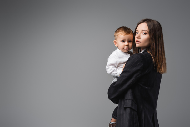 woman in black blazer holding toddler son and looking at camera on grey - Foto, Bild