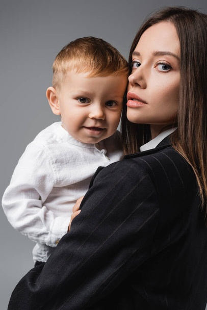 young businesswoman looking at camera while holding little son isolated on grey - Fotografie, Obrázek