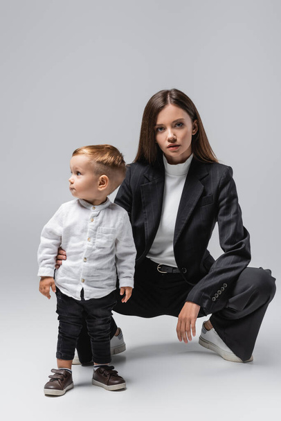 young woman in black suit looking at camera while posing near toddler son on grey - Fotografie, Obrázek