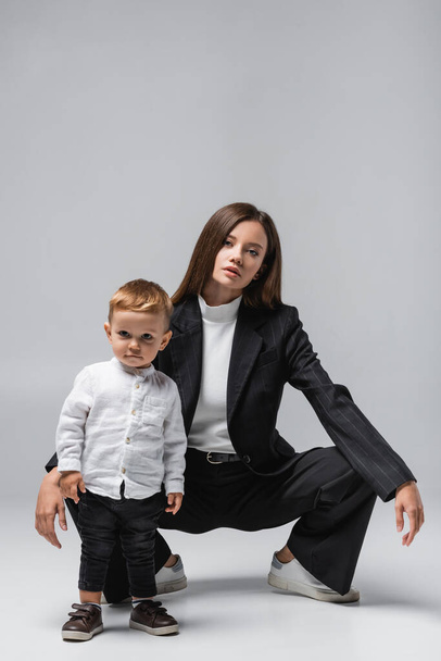 молода жінка в чорному костюмі позує біля маленького хлопчика на сірому
 - Фото, зображення