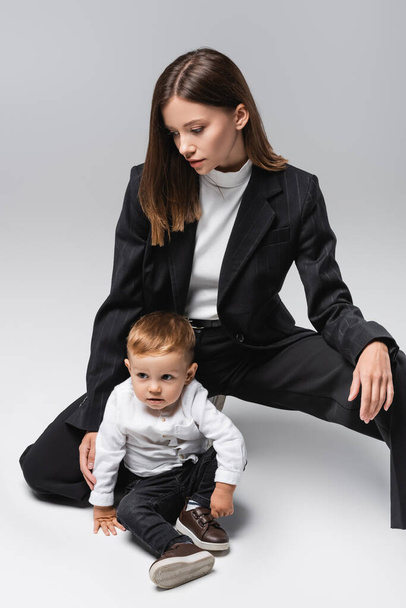 toddler boy sitting near young mom in black suit on grey - Foto, imagen