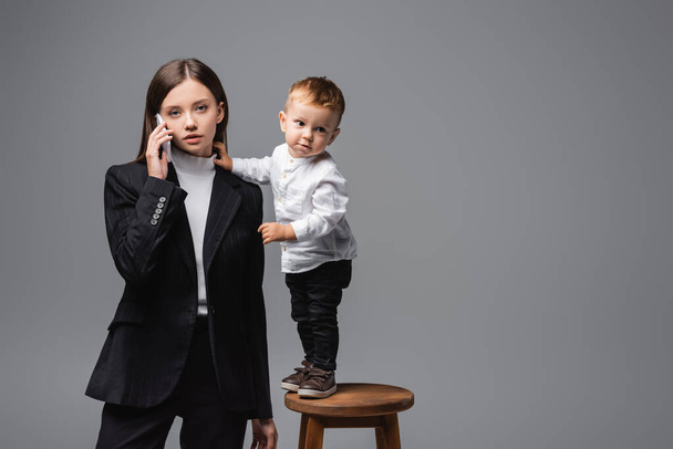 woman in black suit talking on smartphone near little son standing on high stool isolated on grey - Foto, immagini