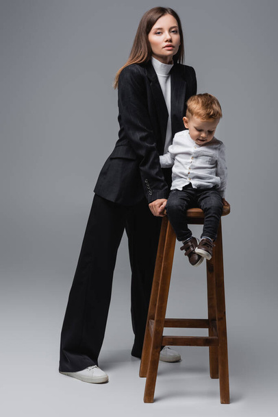 full length view of woman in black suit standing near son on high stool on grey - Φωτογραφία, εικόνα