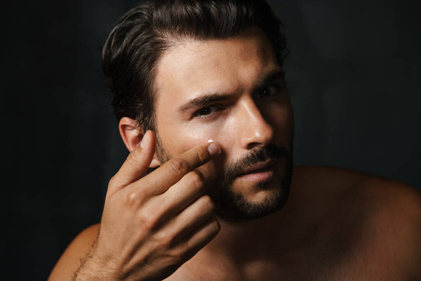 Young half-naked man looking at camera while applying face cream isolated over black background - Photo, image