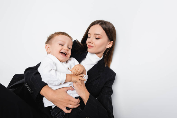 top view of smiling woman in black suit holding cheerful son on white - Zdjęcie, obraz