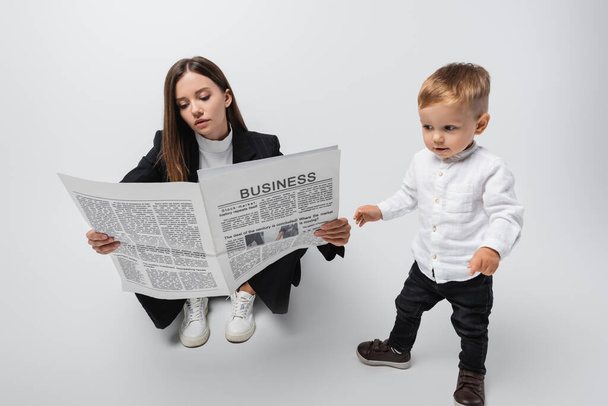 businesswoman sitting and reading newspaper near toddler son on grey - Фото, зображення