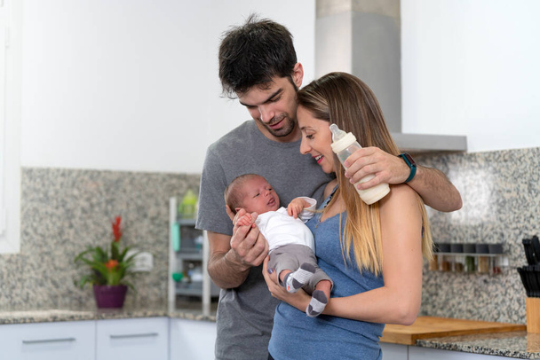 mother and father bottle feeding their newborn baby in the kitchen - Foto, Imagen