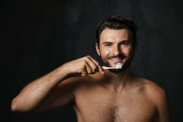 Young half-naked man smiling while brushing his teeth isolated over black background - Photo, image