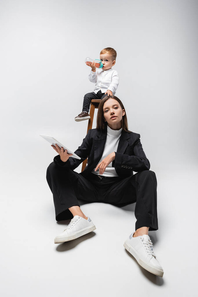 woman in black suit sitting with digital tablet near son on high stool drinking from baby bottle on grey - Fotografie, Obrázek