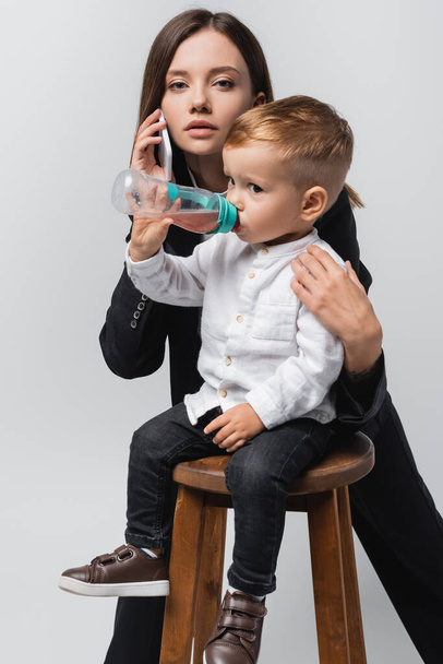 νεαρή γυναίκα μιλάει στο smartphone κοντά στο γιο πόσιμο από το μωρό μπουκάλι απομονωμένο σε γκρι - Φωτογραφία, εικόνα