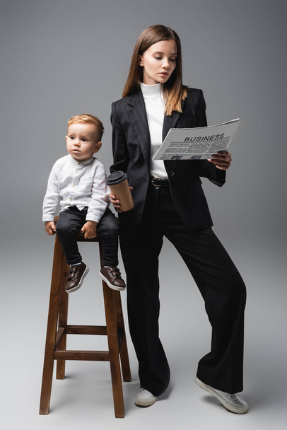toddler boy sitting on high stool near mother with coffee to go and business newspaper on grey - Foto, immagini