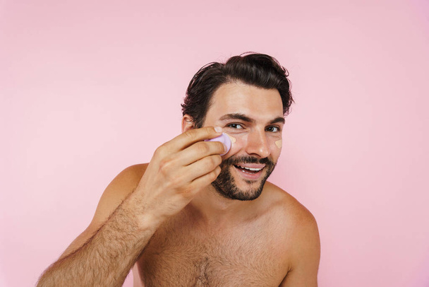 White shirtless man smiling while applying concealer with sponge isolated over pink background - Fotoğraf, Görsel